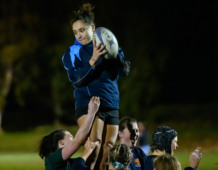 A group of women play rugby on an outdoors pitch at night.
