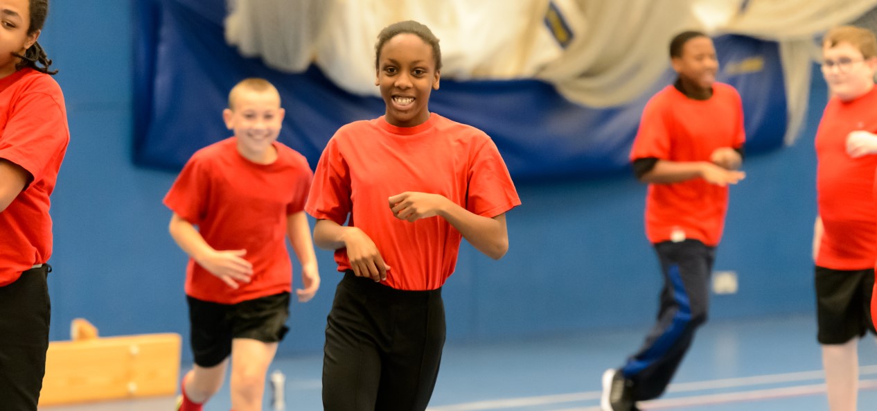A schoolgirl smiles while jogging among fellow pupils in a sports hall.