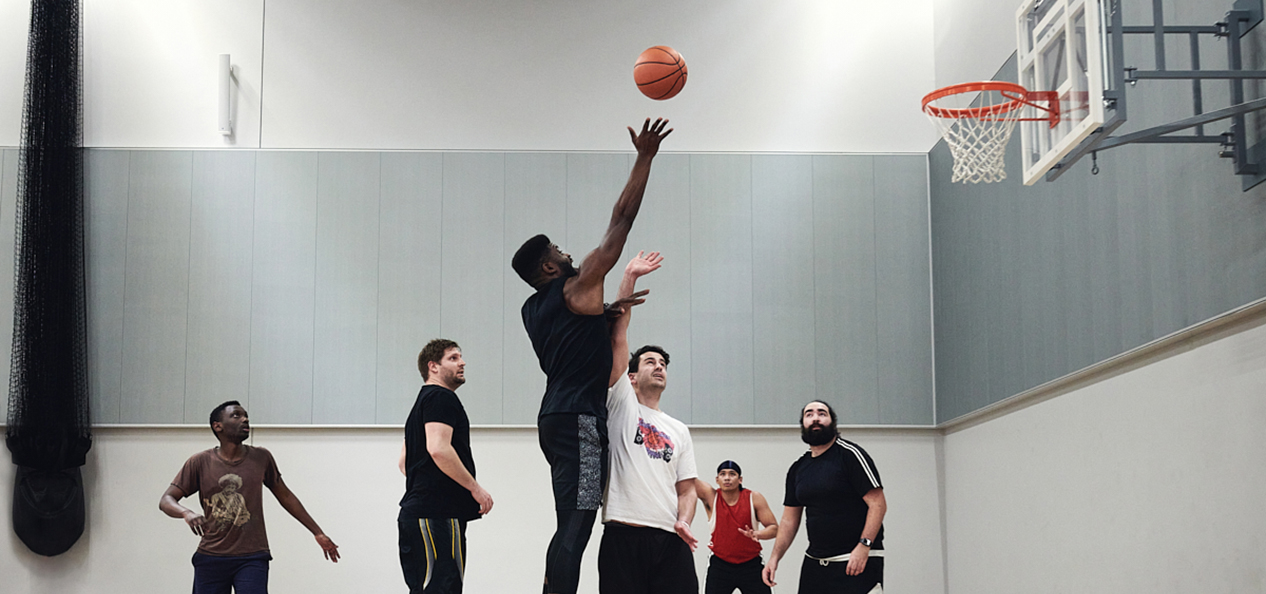A man shoots a basketball over another player who tries to block it, while other players watch on
