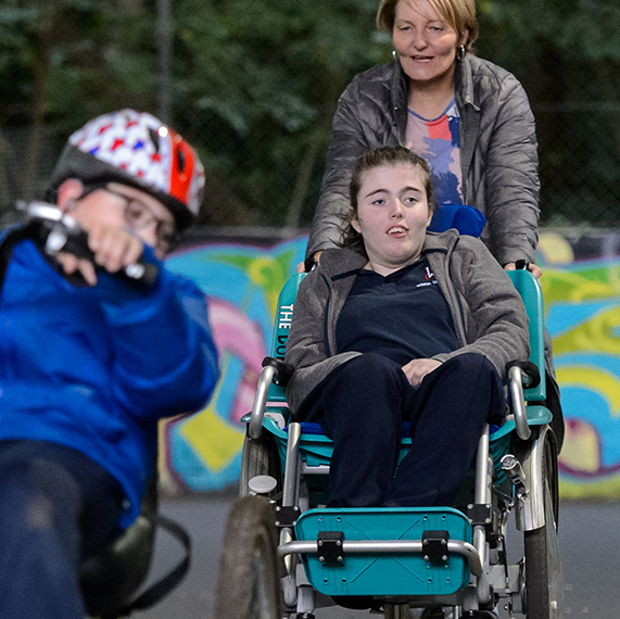 A girl on chair with big wheels is pushed by her mother, while a boy wearing a helmet cycles ahead of them.   