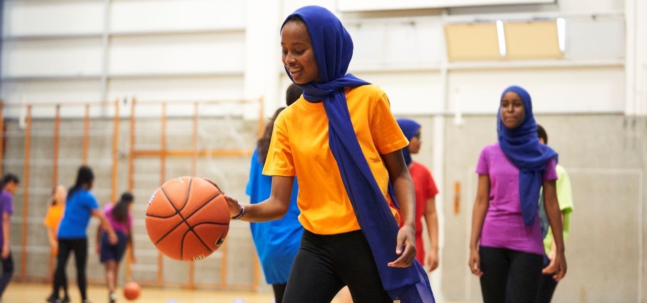 A girl in a headscarf is on a basketball court - she's smiling and about to bounce the ball