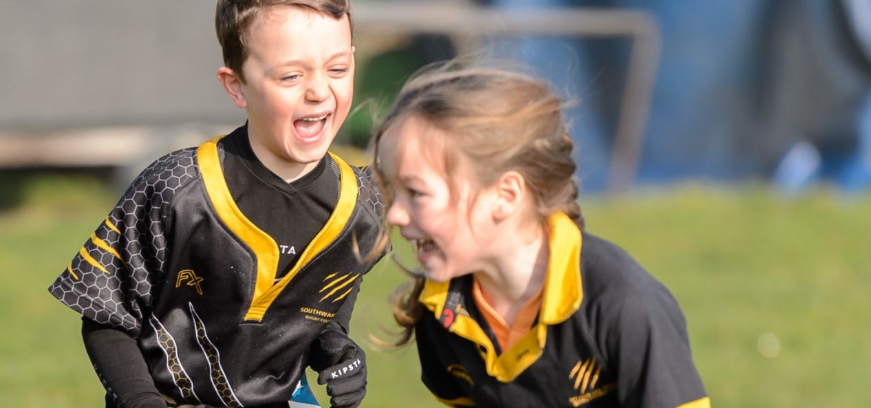 A young boy chases a girl during a game of tag rugby, with both players sporting wide smiles.