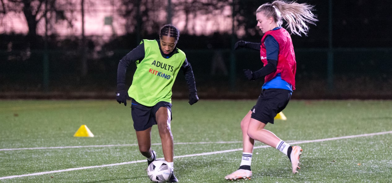 A female footballer looks to dribble away from her opponent during a game on an outdoor artificial pitch.