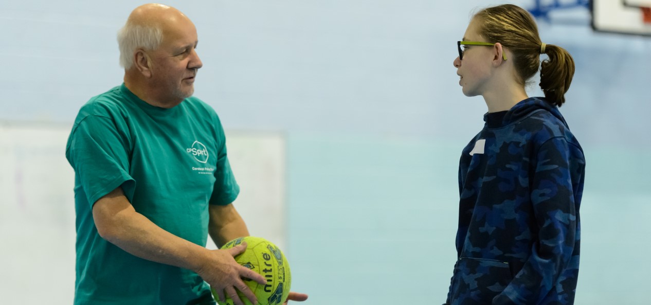 An older man holding a football talks to a girl in a sports hall.
