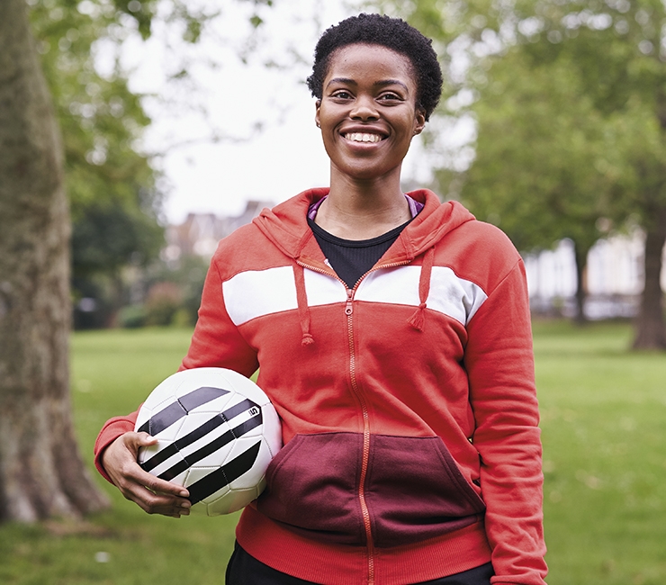 A black woman smiles while holding a football in the park.