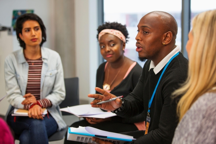 A man holding a pen and notepad talks to a group of people sat around him in an office.