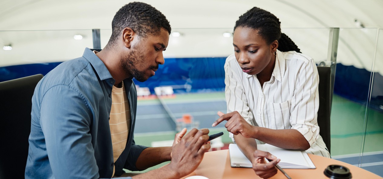 Two people consider something on a mobile phone while sat at a work desk above indoor tennis courts.