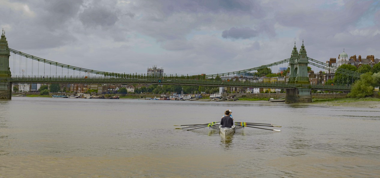 Three people in a rowing boat head towards a bridge on the River Thames.