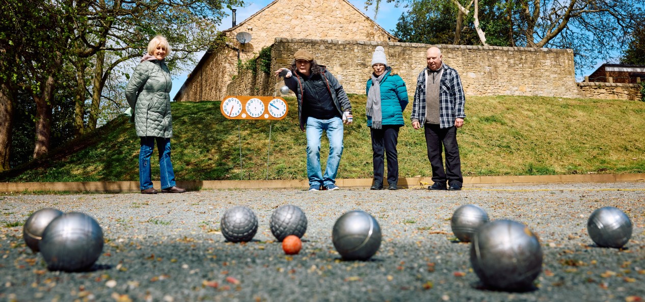 An older woman throws a ball during a game of petanque, while three fellow players watch on.