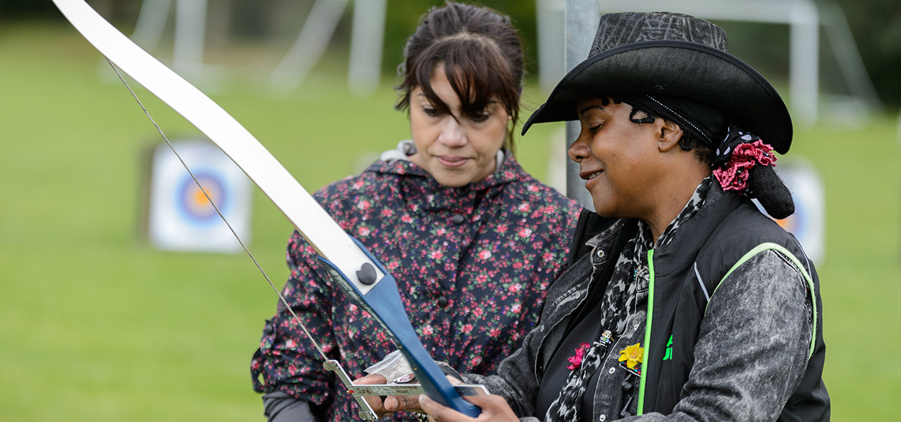 A female archery coach instructs another woman at an archery lesson outdoors.