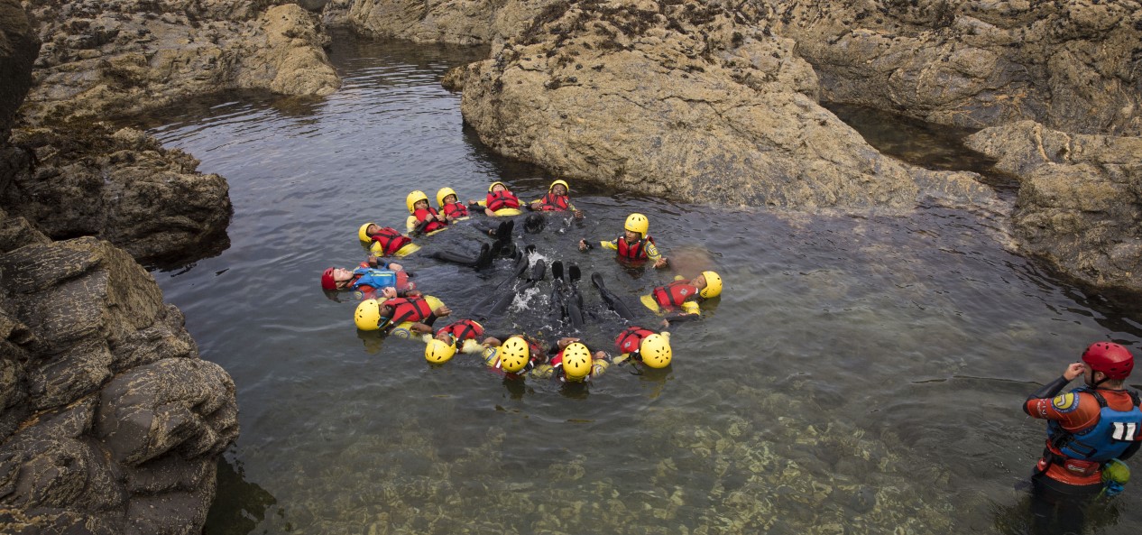 A group of people wearing helmets and buoyancy vests scull in a circle in some shallow sea water, surrounded by rocks.