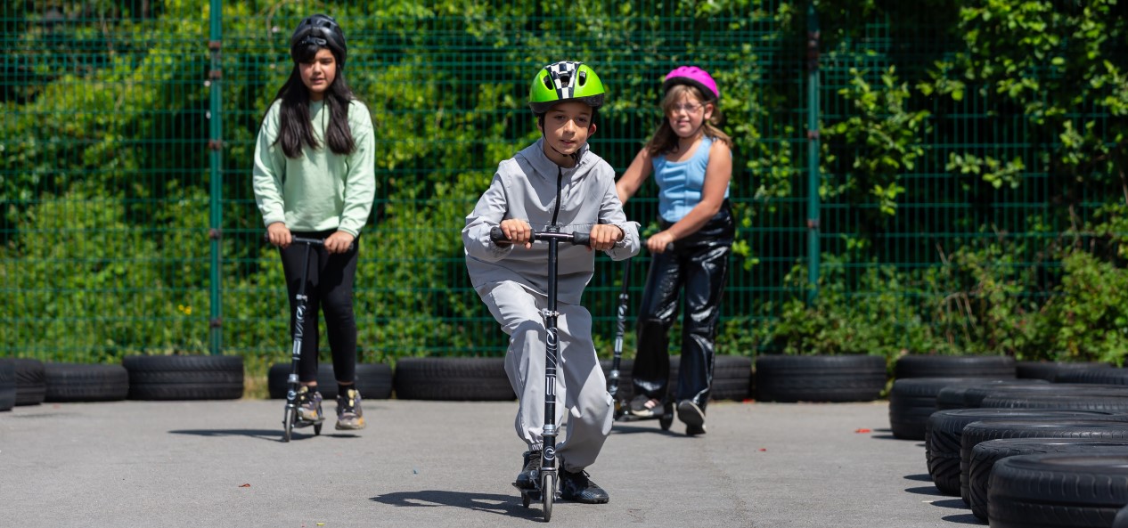 Three children wearing helmets ride on scooters.