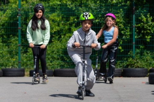 Three children wearing helmets ride on scooters.