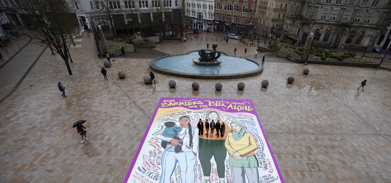 In the centre of Birmingham, five women stand on a giant mural featuring illustrations of women and the text 'Some barriers are too big to tackle alone'.